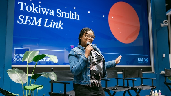 Black woman with glasses holding a microphone standing in an auditorium.