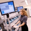 A photograph of Geneticist Pardis Sabeti working in a laboratory. She is standing beside a wheeled set of shelves holding multiple square containers. She is also looking towards a computer monitor sitting close to the shelves and her gloved hands are poised on a device with a big red button.