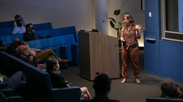 A woman with a pointer presenting slides to a crowd.