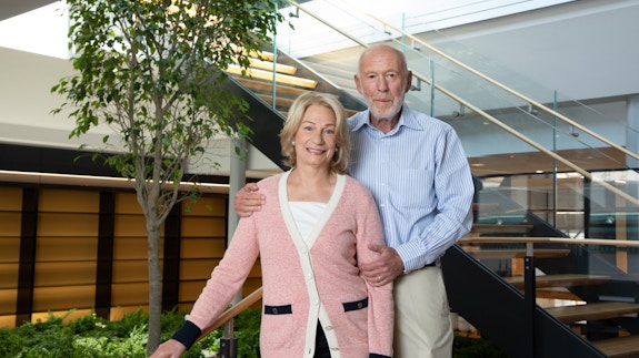A photograph of Marilyn and Jim Simons. Marilyn is a woman with light blonde hair and is wearing a pink, black, and white button sweater with a white undershirt and black slacks. Jim Simons is a man with a faint white beard, a blue-and-white pinstripe button-down shirt, and beige slacks. They are situated in a room with a staircase leading upward and that has a small garden with a tree underneath it.