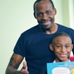 A man standing with this thumbs up behind a young boy with a paper containing the SPARK program logo and the title 