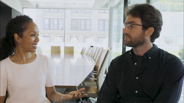 Two people conversing in a business boardroom. One of the people is smiling while the other has a neutral expression.